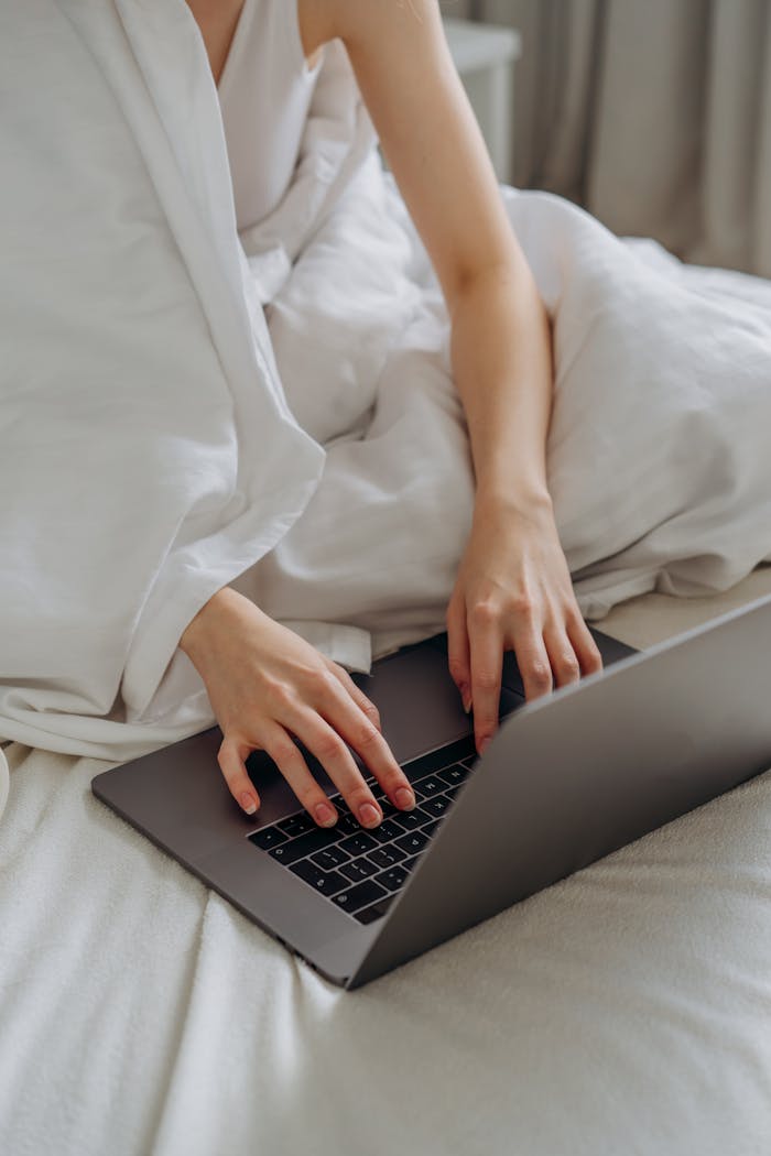 Adult woman comfortably typing on a laptop while sitting in bed covered with white linens.